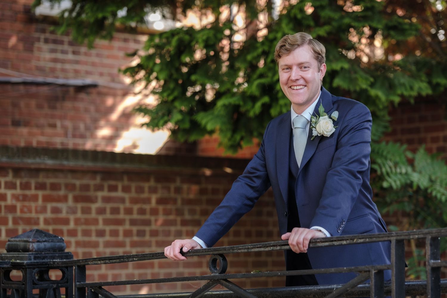 A person in a navy blue suit with boutonniere leans on an outdoor metal railing near a brick wall and trees.