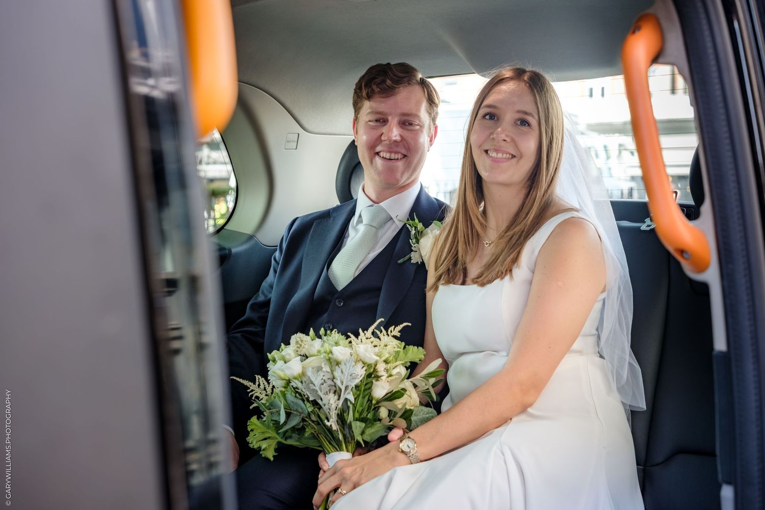 A couple sits in the back of a car with white floral bouquet and formal wedding attire.