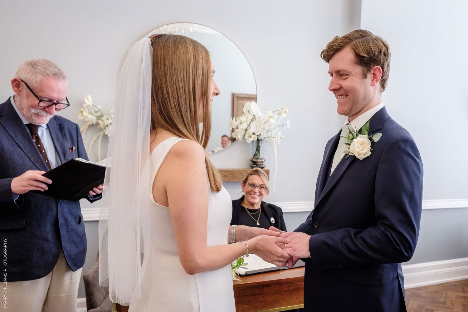 Bride and groom exchange rings during an intimate indoor wedding ceremony with an officiant present.