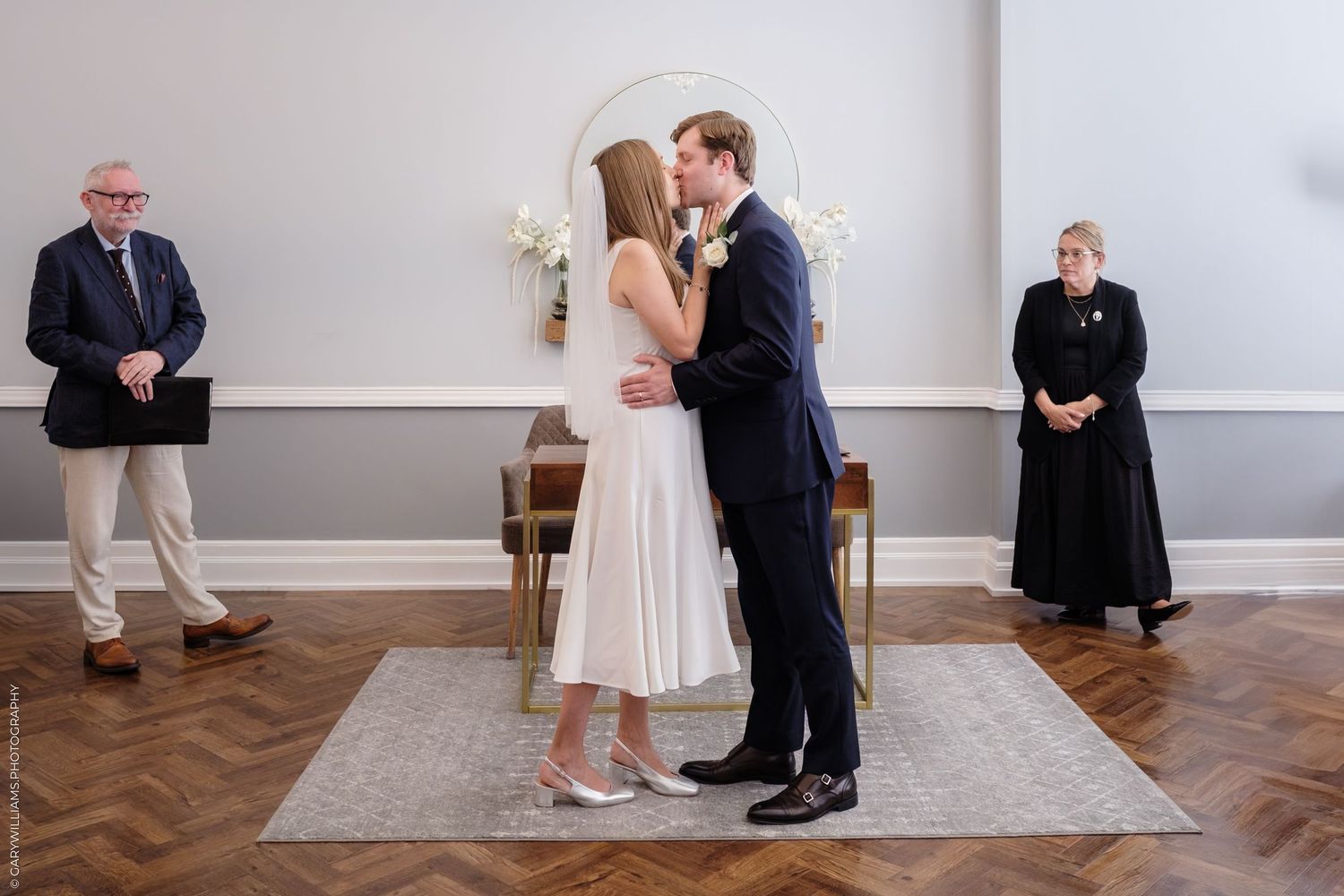 A couple shares their first kiss during an intimate indoor wedding ceremony while two officiants look on.