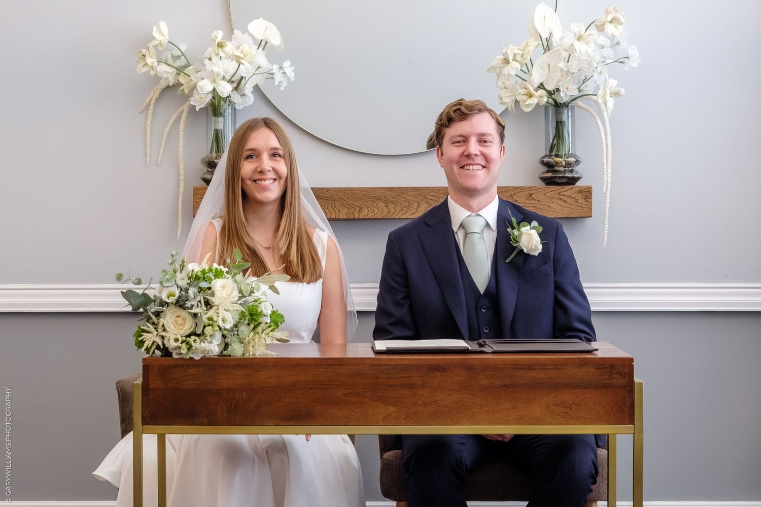 A newlywed couple signing papers at a wooden desk decorated with white floral arrangements and a round mirror.