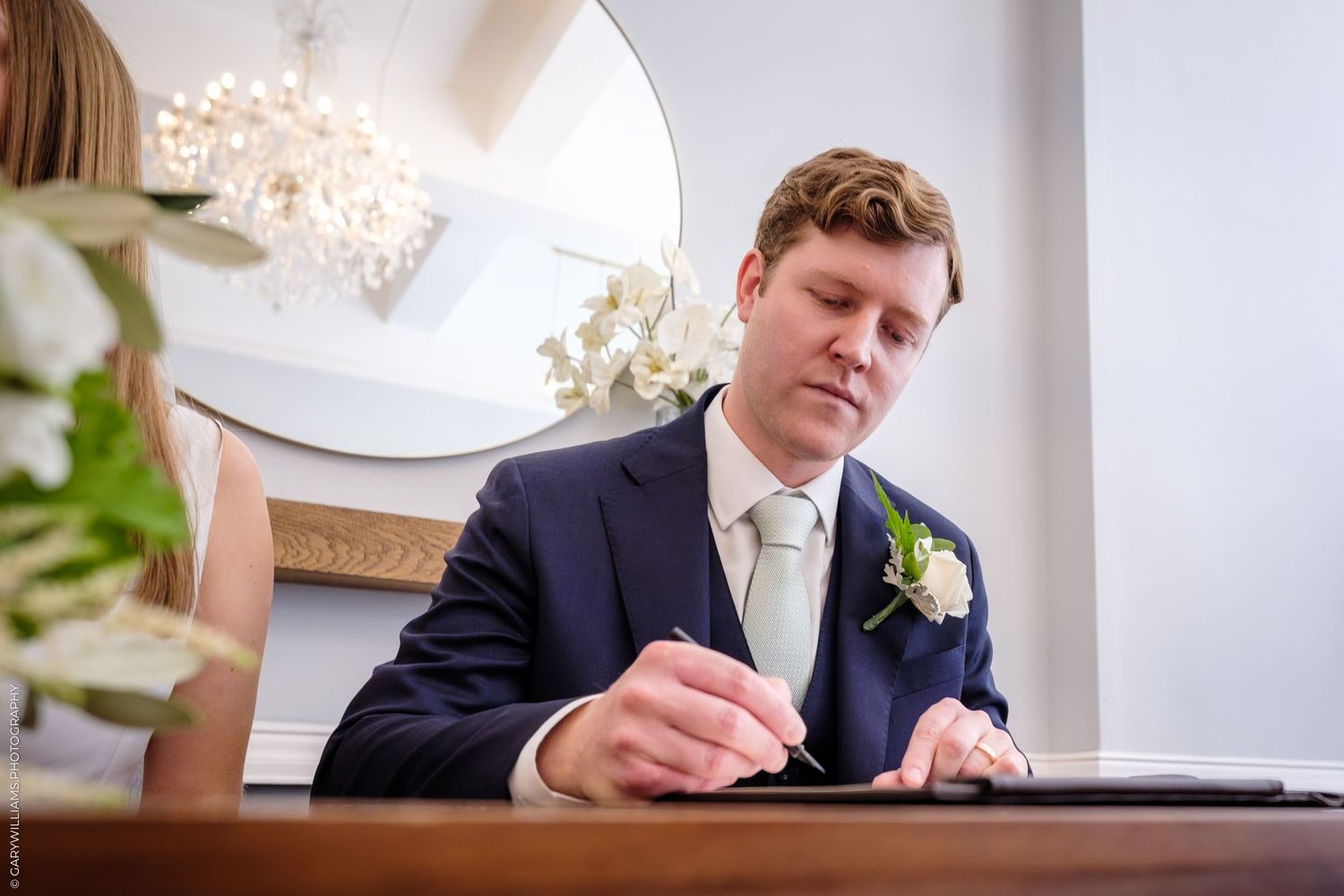 A person in a navy suit signs a formal document at a table with a coffee cup.