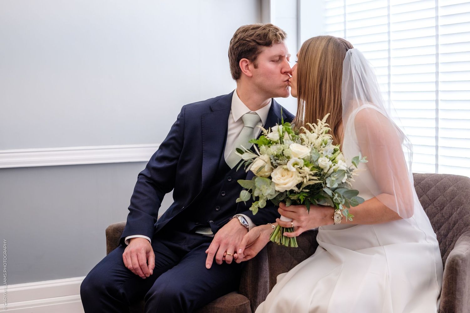 A couple shares a tender moment while seated, with the bride holding a white floral bouquet.