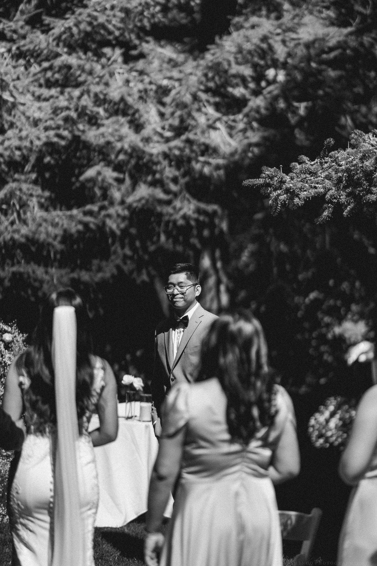 A black and white photograph capturing a candid moment during an outdoor wedding ceremony.