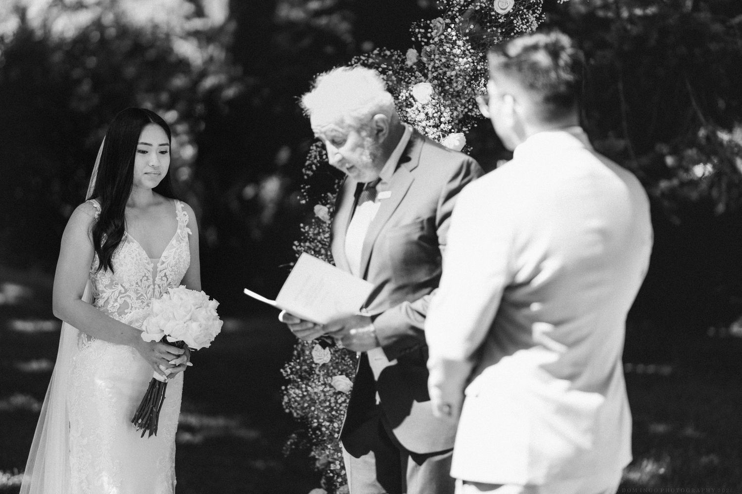 A black and white photograph capturing an intimate outdoor wedding ceremony with an officiant and couple under trees.