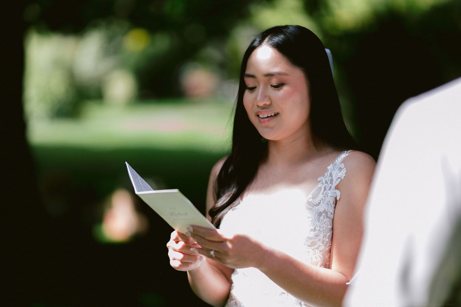 Woman in white lace dress reads card while sitting outdoors in garden setting.