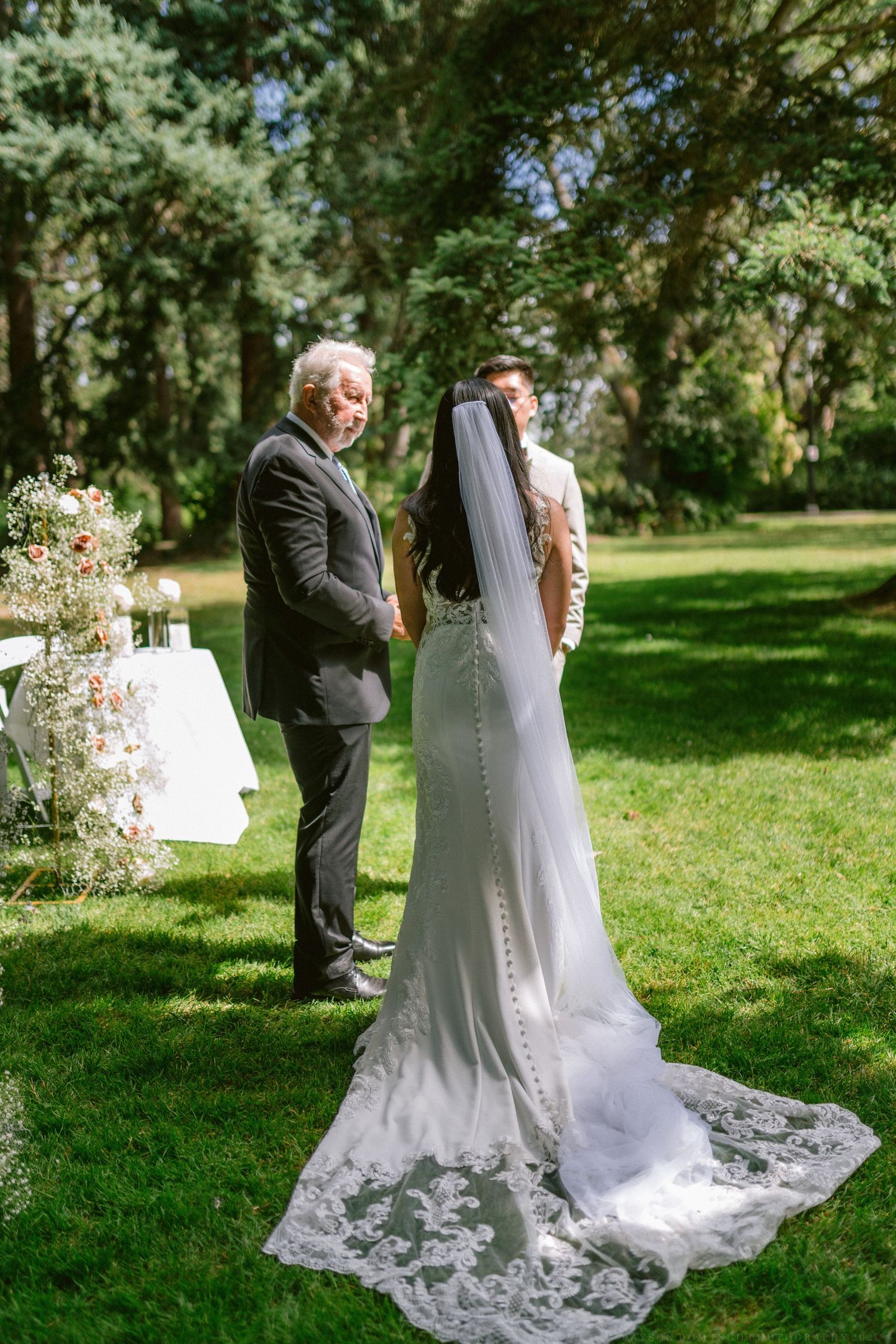 Bride in flowing lace train gown shares a moment with a guest in a garden setting wedding ceremony.