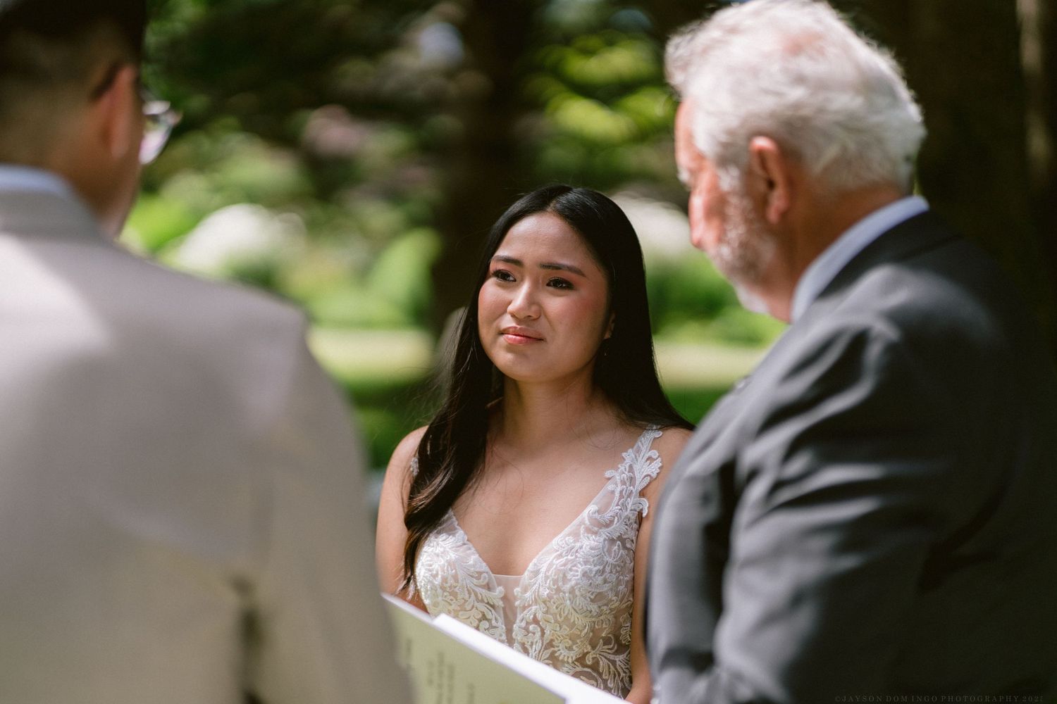 A bride in a lace wedding dress listens intently during an outdoor wedding ceremony.
