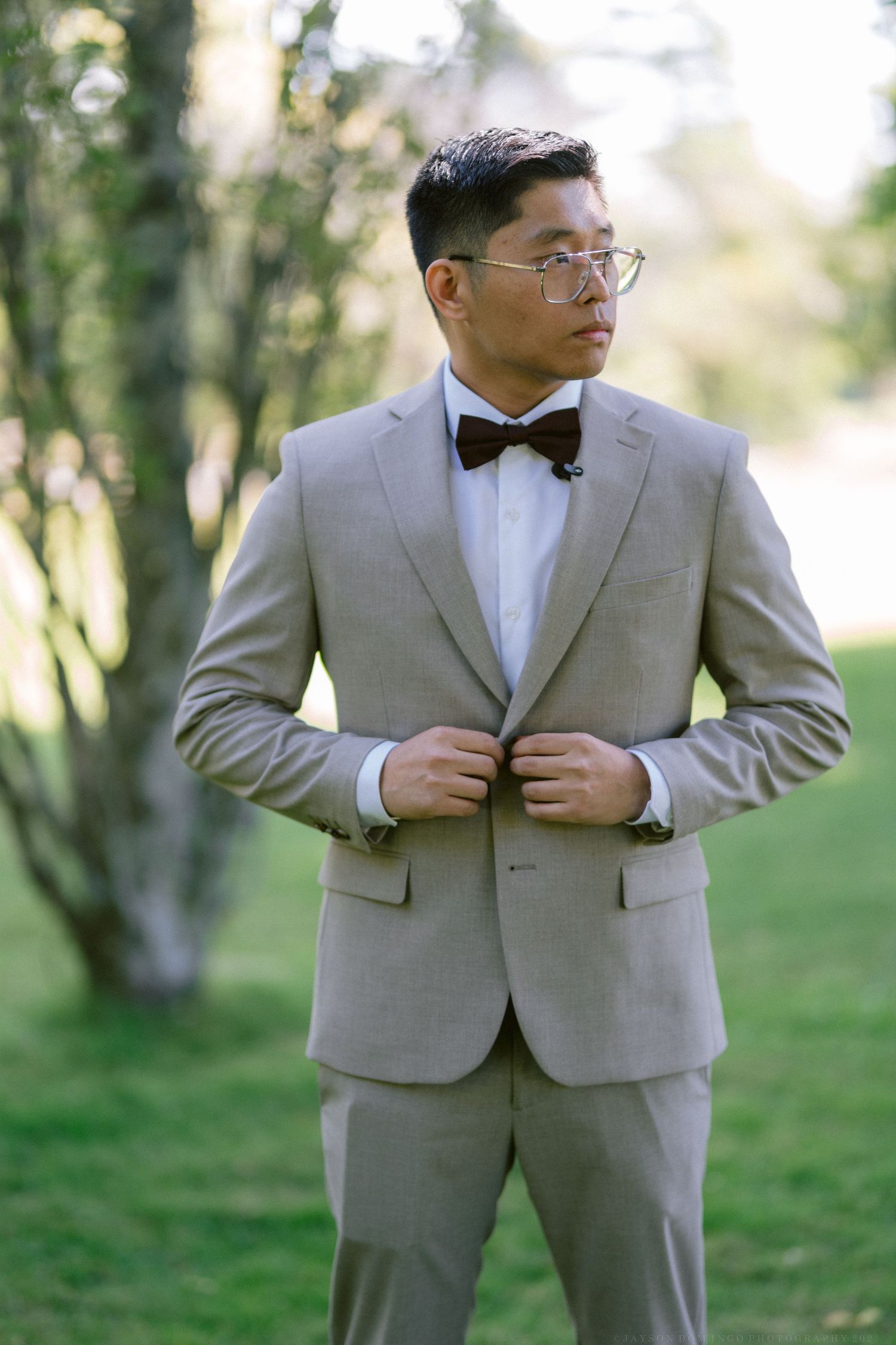 A person in a light gray suit and black bow tie poses outdoors in a garden setting.