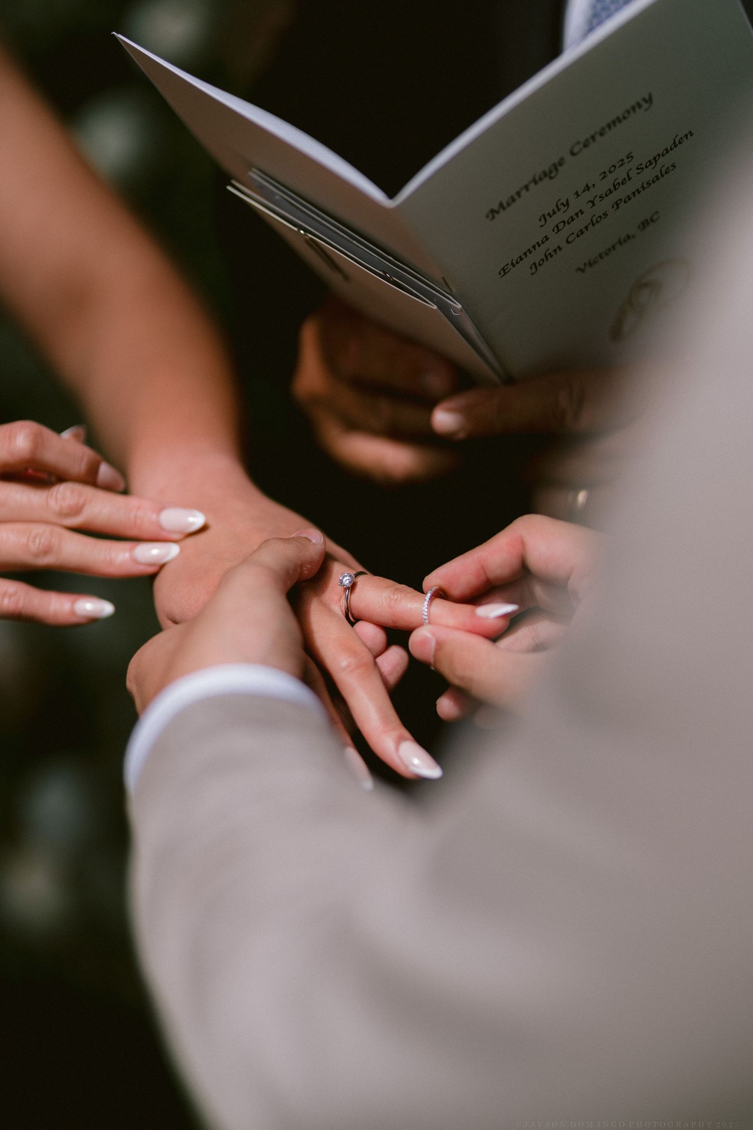 Close-up view of hands exchanging wedding rings during marriage ceremony with blurred book background.