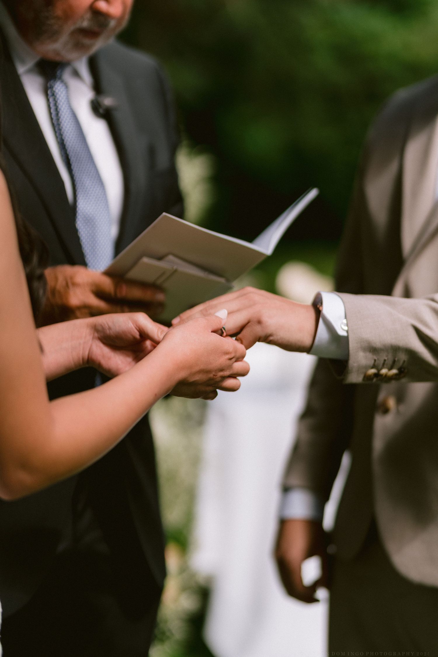 A close-up shot of hands exchanging rings during an outdoor wedding ceremony.