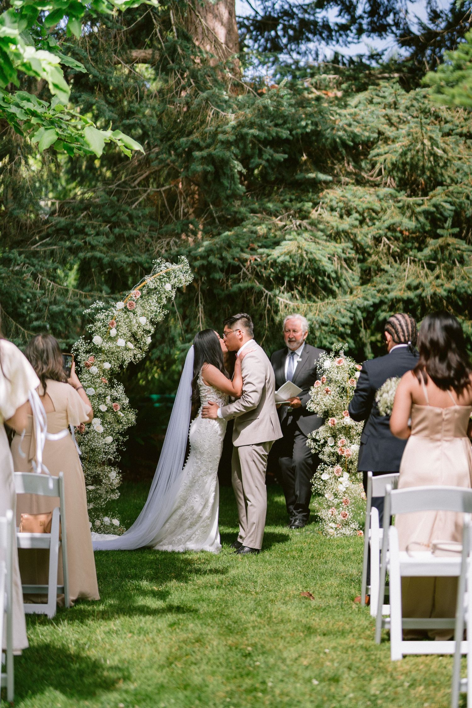 An outdoor garden wedding ceremony takes place on a lush green lawn surrounded by tall trees and white floral decorations.
