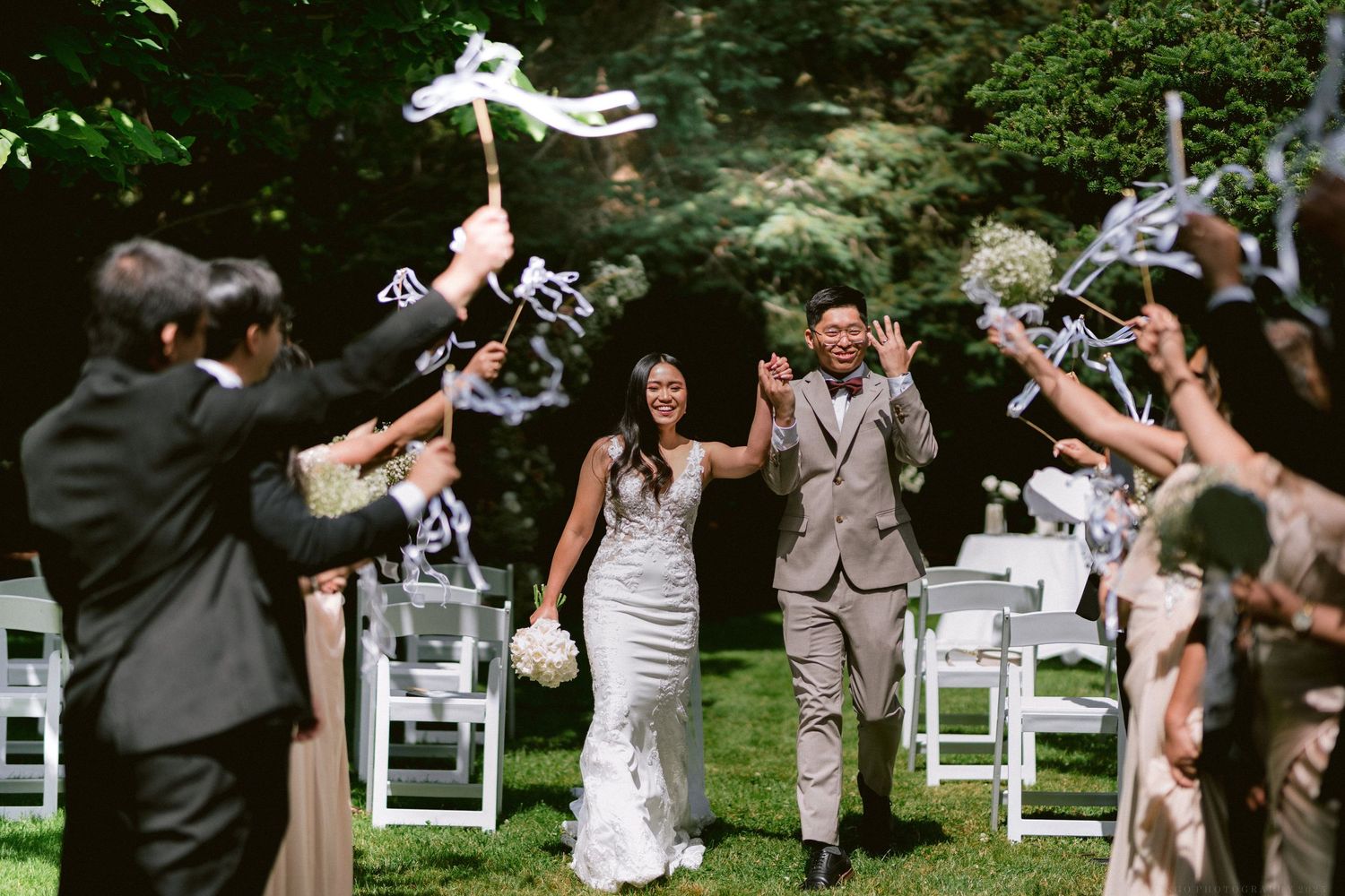 Joyful wedding party wearing grey and white formal attire celebrates with confetti outdoors in a garden setting.