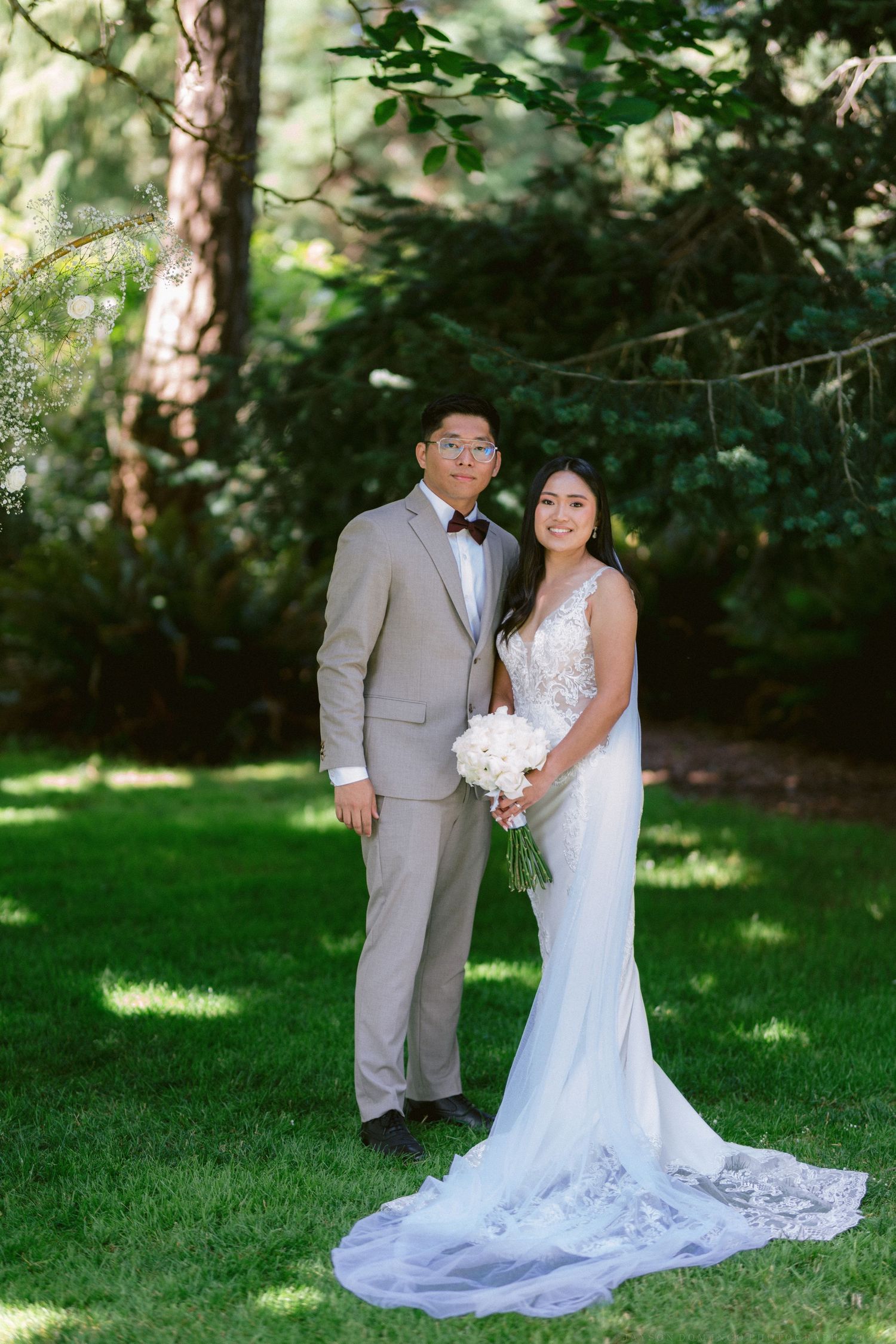 A couple in wedding attire poses in a garden setting with lush greenery and trees in the background.