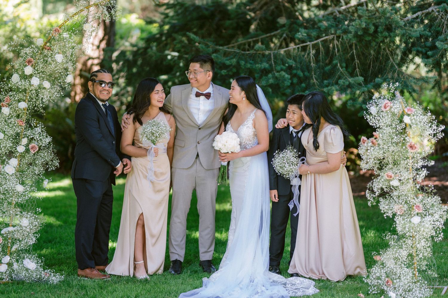 A bridal party gathered outdoors surrounded by blooming white flowers and greenery.