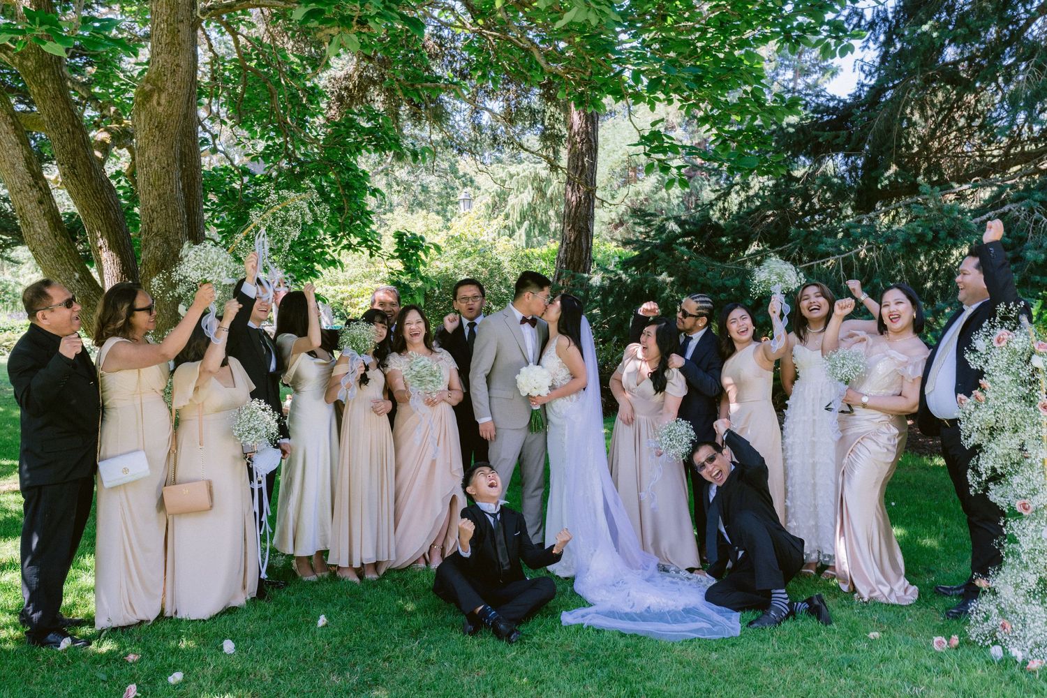 Wedding party poses together in coordinated blush and neutral dresses under lush garden trees.