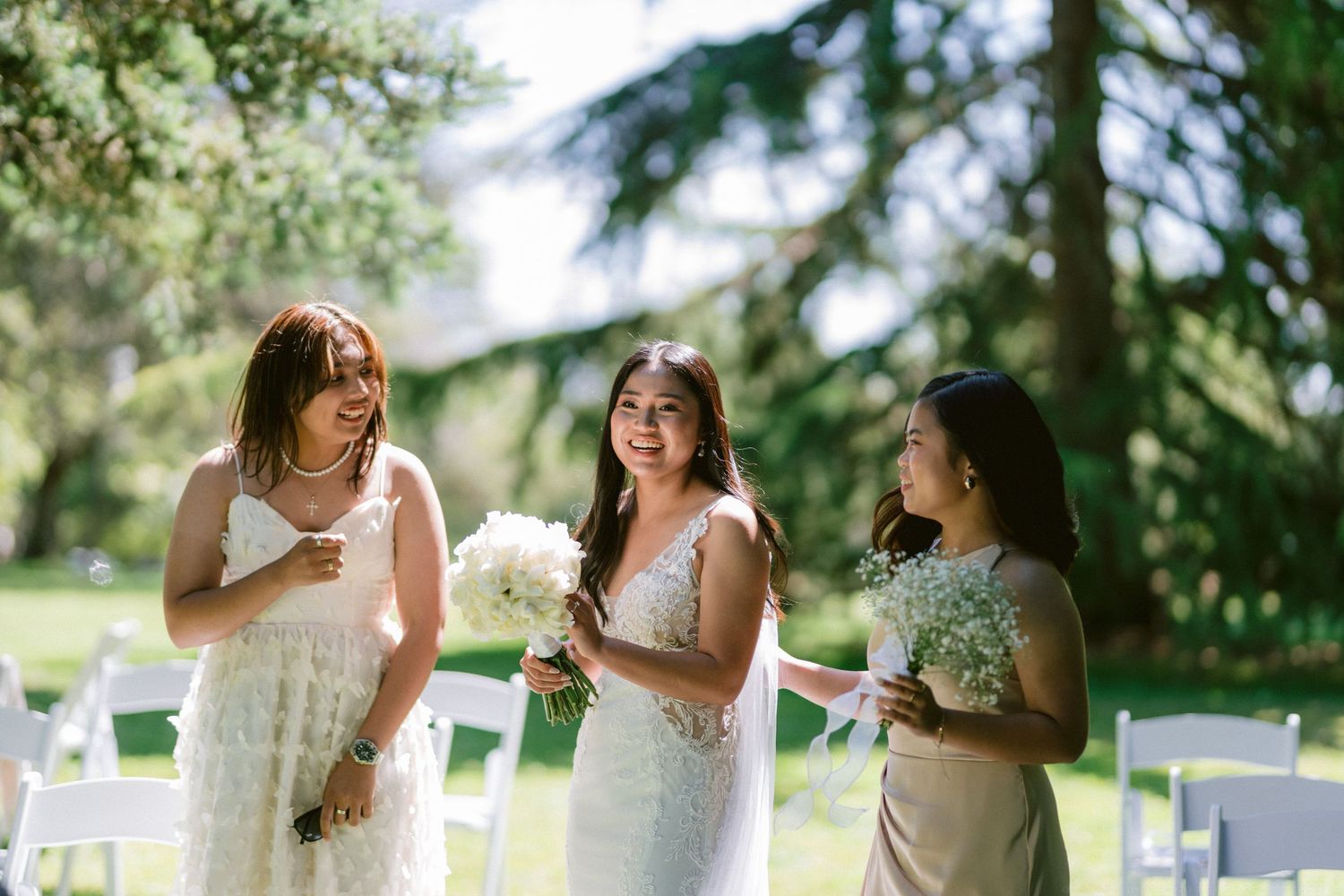 Three women in white dresses share a candid moment during an outdoor wedding celebration.