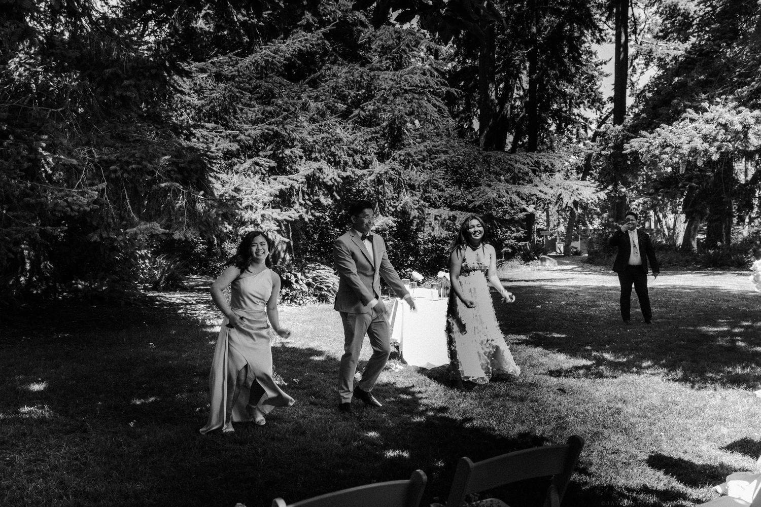 A black and white photograph of a wedding party walking through a tree-lined path.