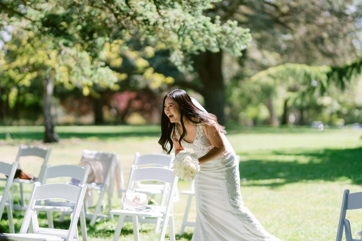 A white wedding dress being adjusted among rows of white folding chairs on a sunny lawn.