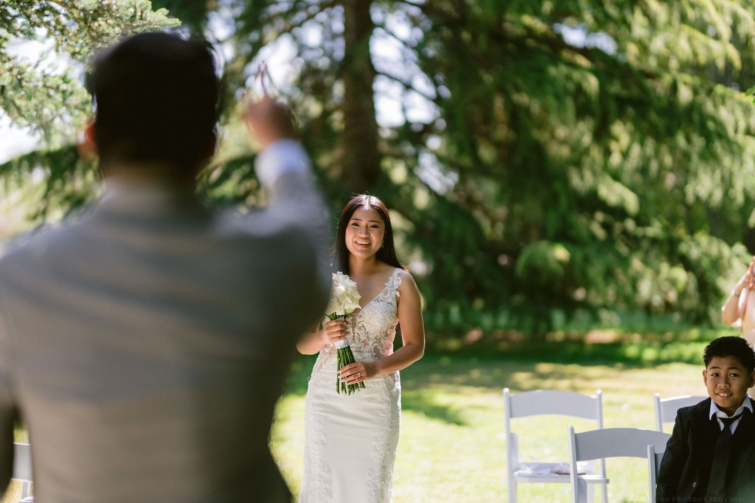 A garden wedding ceremony with white chairs arranged beneath towering evergreen trees.
