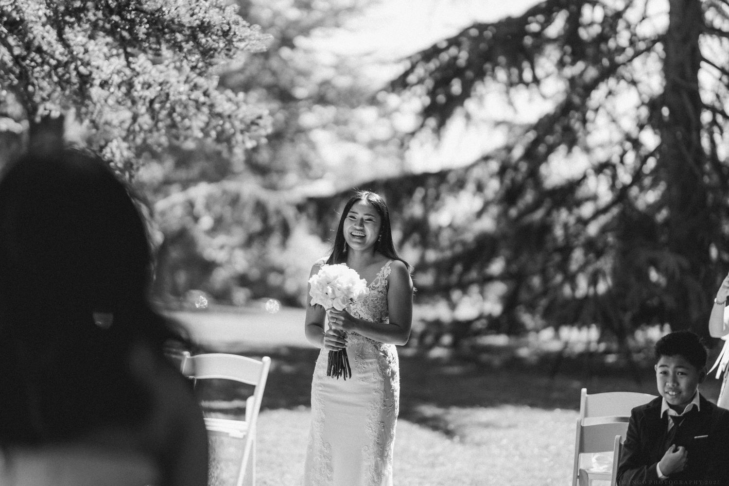 A wedding ceremony outdoors with many white chairs arranged on grass under trees.