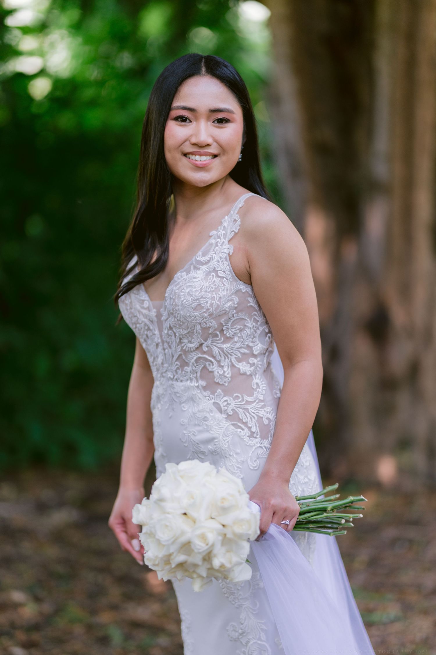 A glowing bride in an elegant lace wedding gown poses with a classic white rose bouquet in a romantic forest setting.