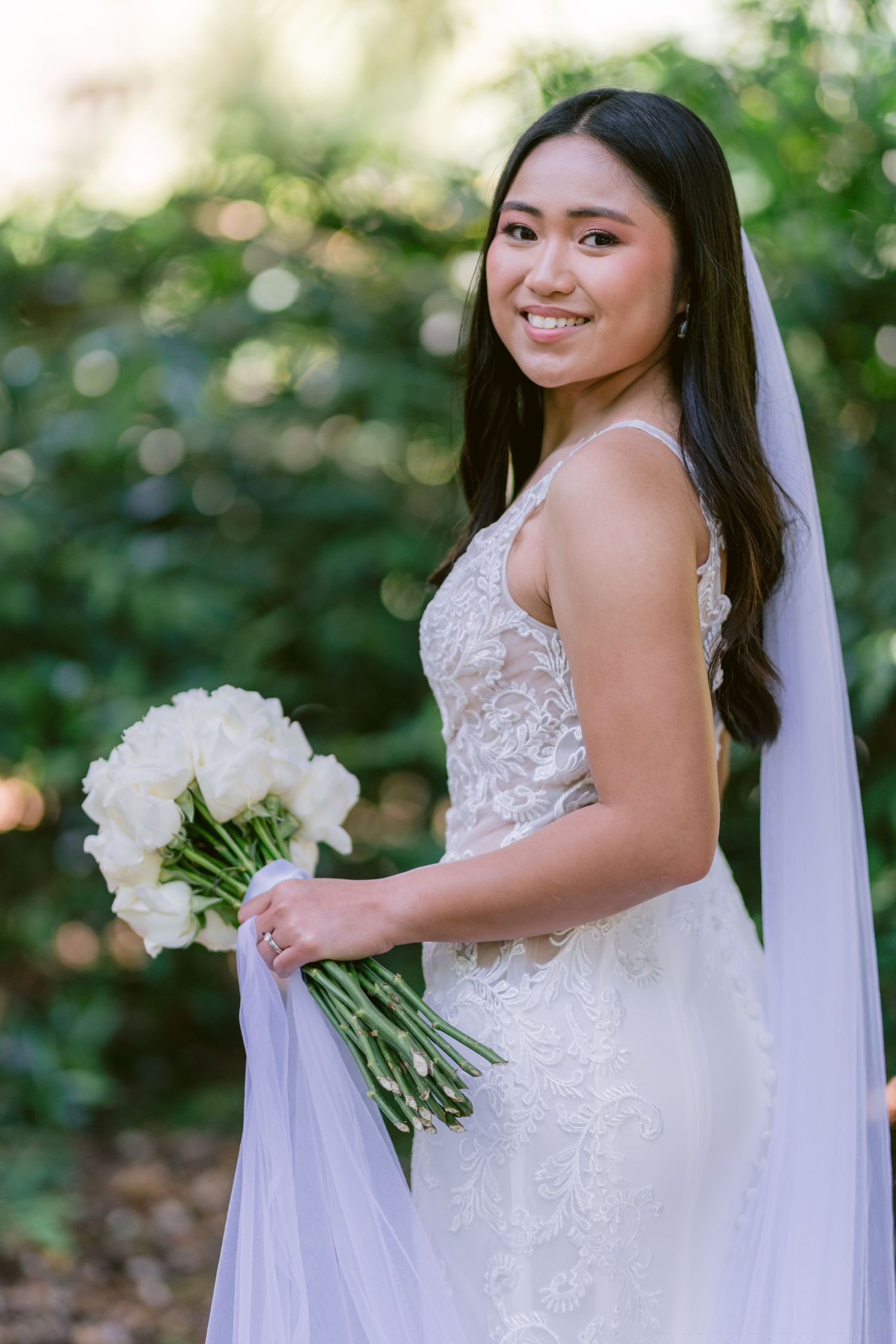 A bride in a lace wedding dress holds a white bouquet while wearing a flowing veil in an outdoor garden setting.