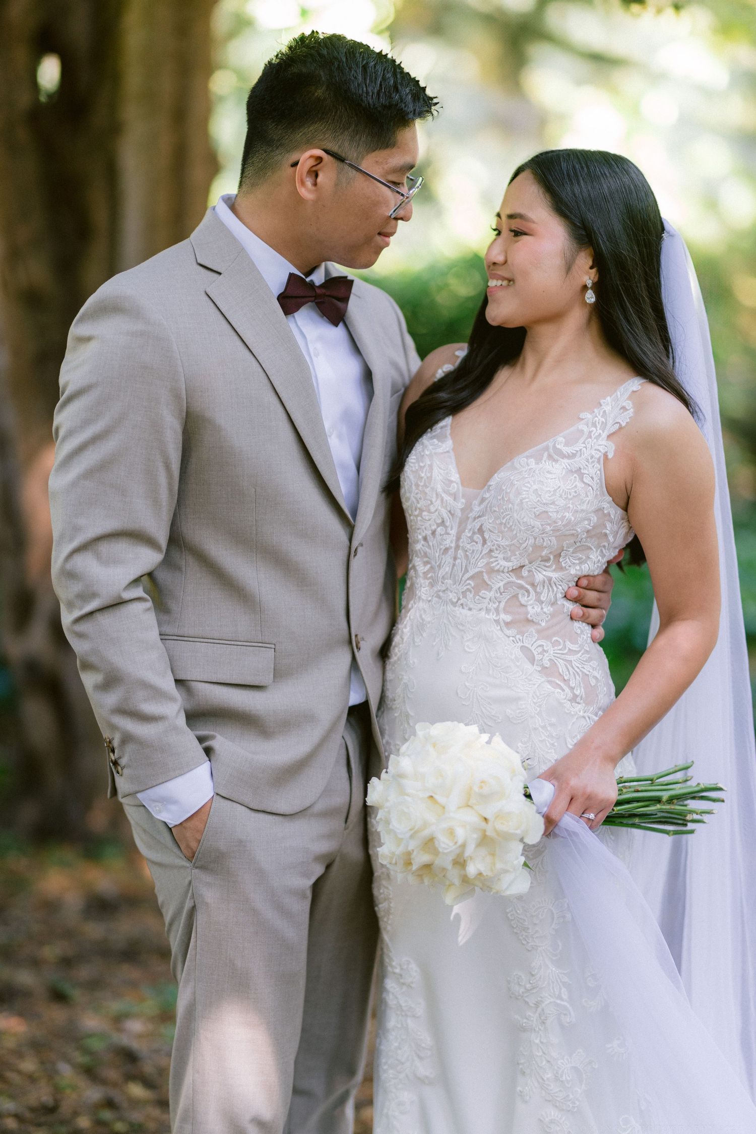 A wedding couple poses together in formal attire under trees with white bouquet.