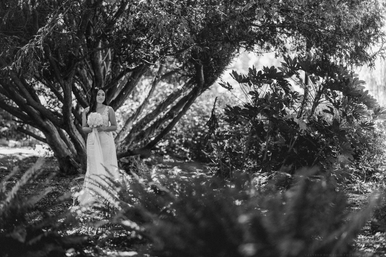 A dreamy black and white photo of a bride in a garden setting with sunlight filtering through trees.