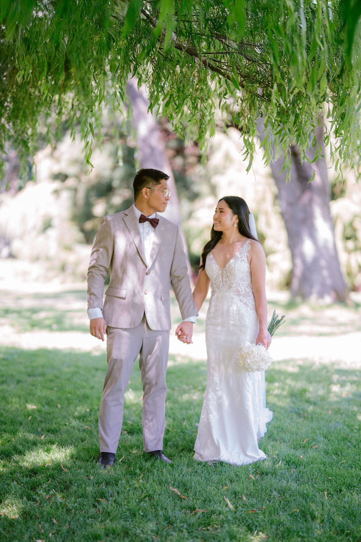 Couple in wedding attire walking together under weeping willow trees.