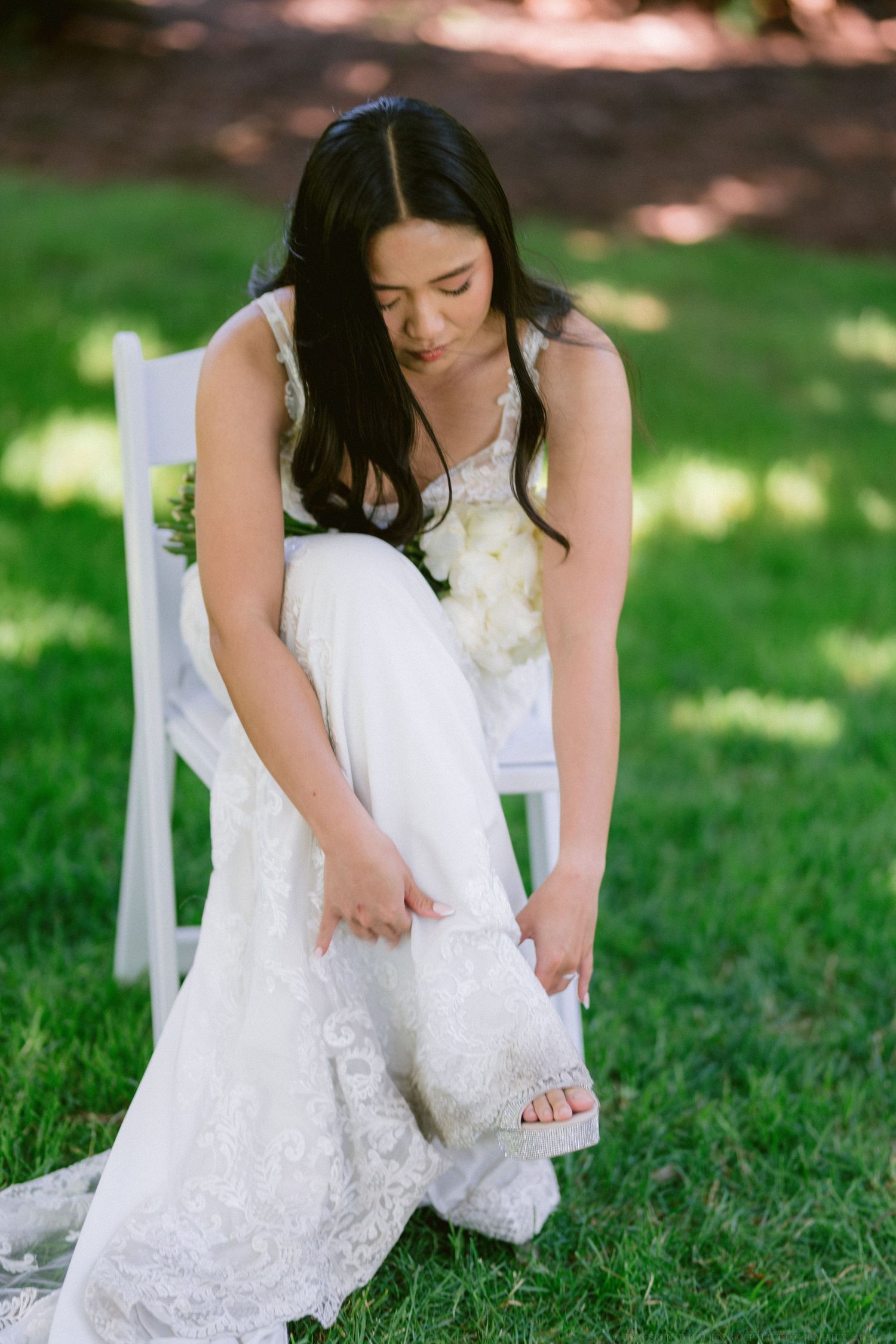 A bride in a white lace gown adjusts her shoe while sitting on a white chair on a grass lawn.