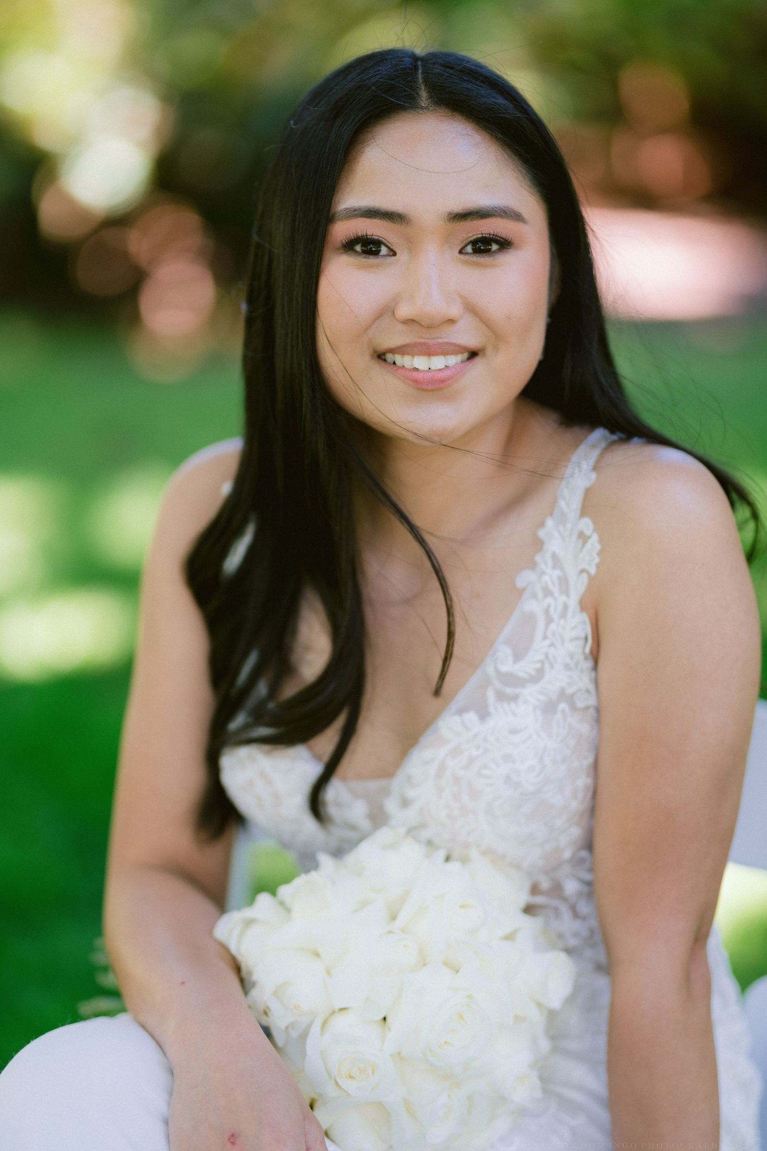 A bride in a lace wedding dress holds a bouquet of white roses in a garden setting.