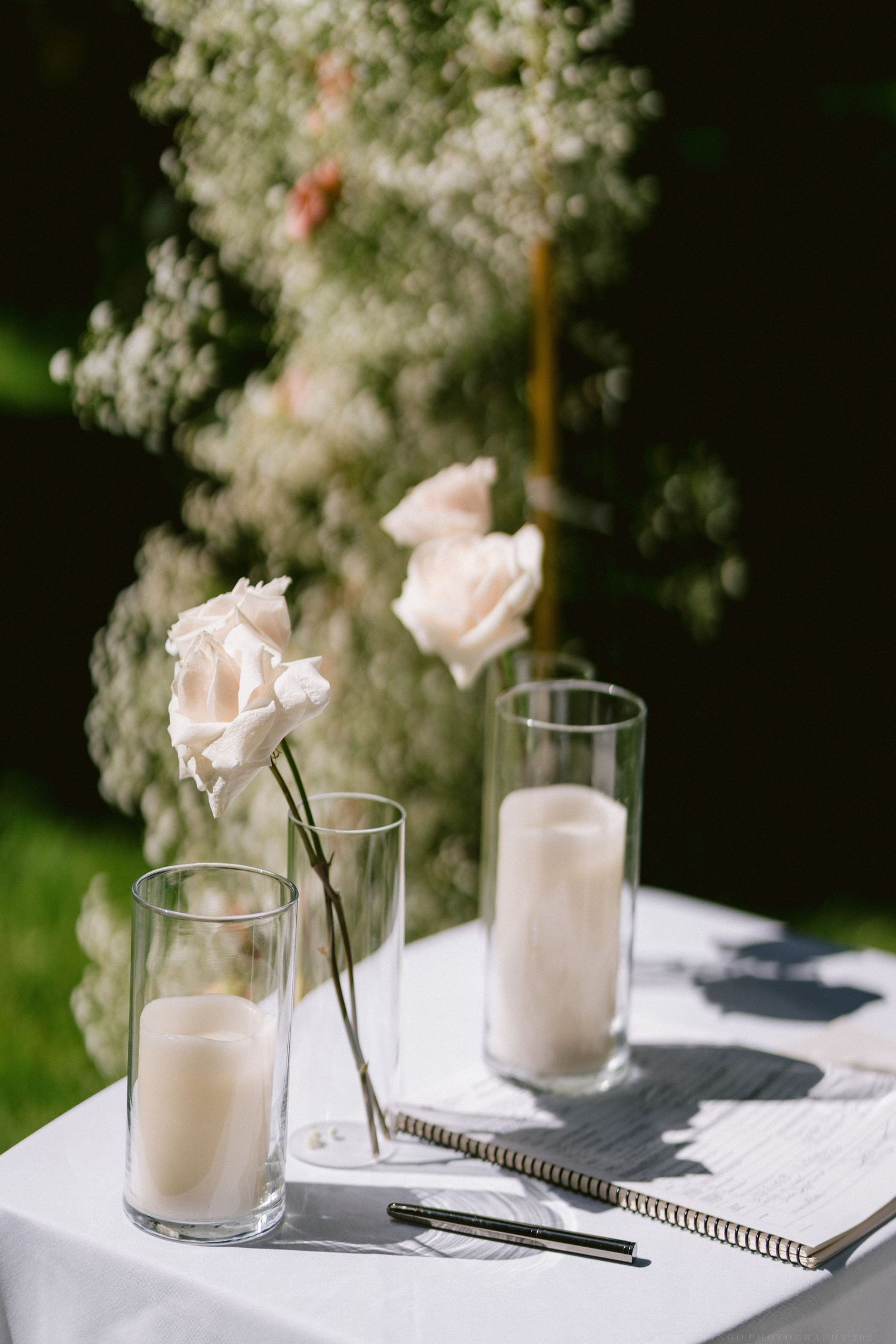 Pink roses in glass vases with milky liquid alongside a guest book and pen on a white tablecloth.