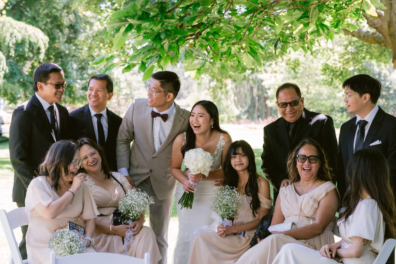 A group enjoying candid moments during a wedding celebration under green trees on a sunny day.