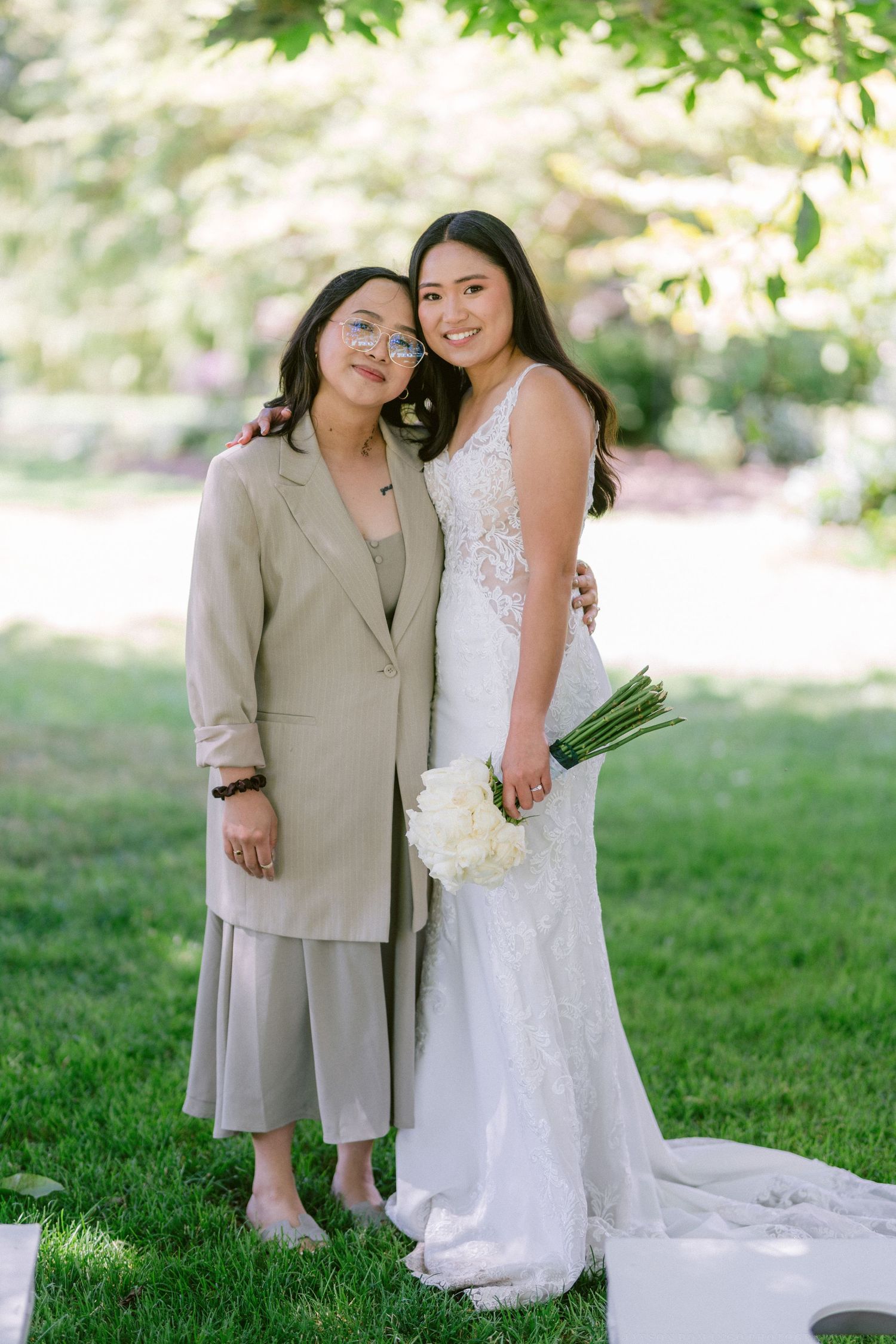 Two elegantly dressed wedding guests embrace in a sunny garden setting.