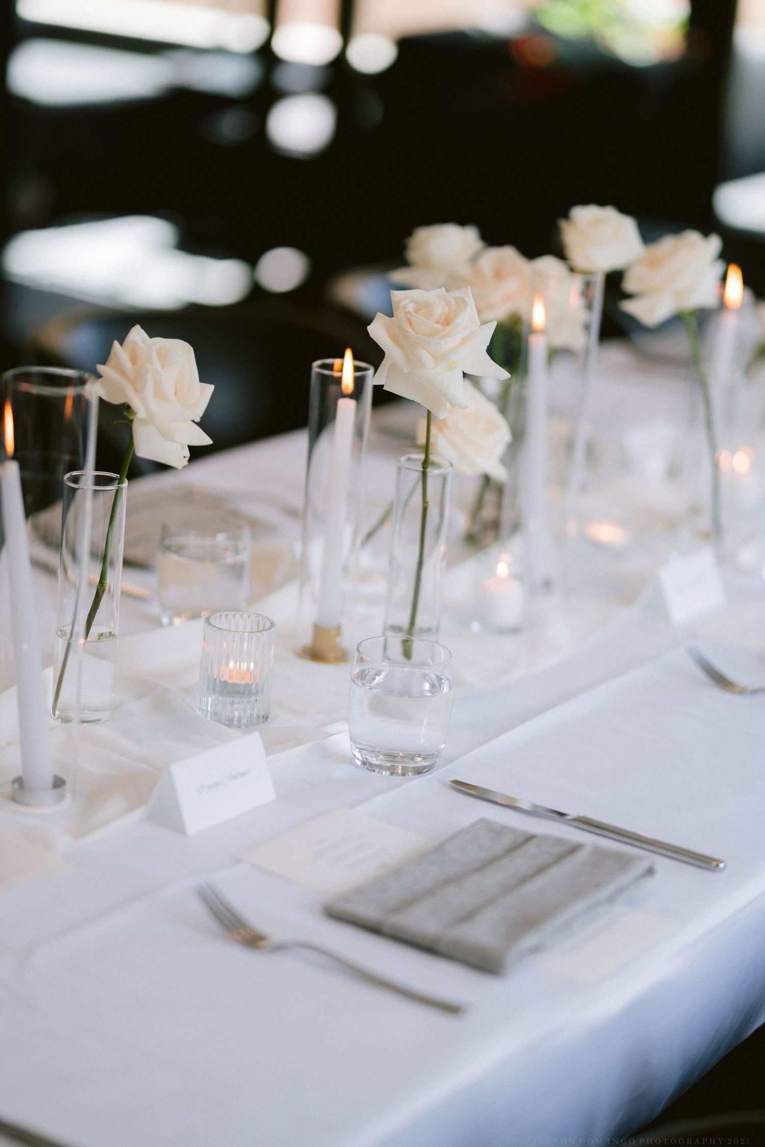 Elegant table setting with single white roses in tall vases and white linens.