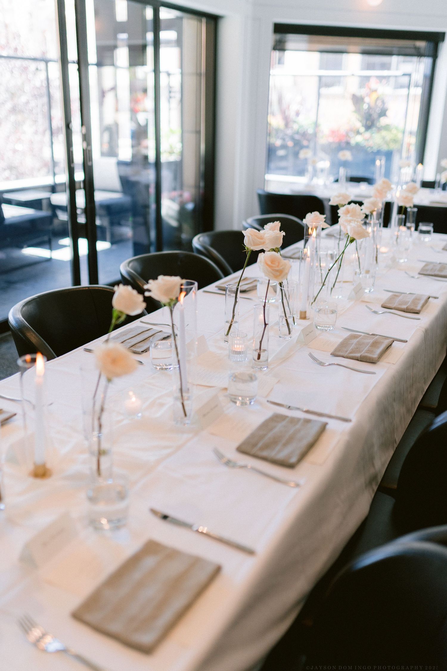 Modern restaurant table setting with white roses in bud vases and natural lighting.