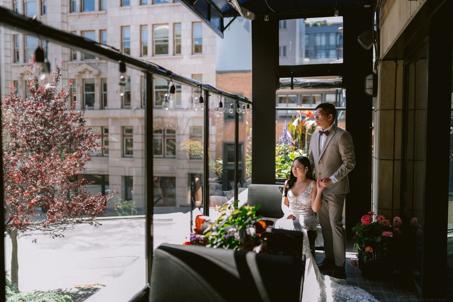 A couple stands on an urban balcony with cherry blossoms and city buildings in view.