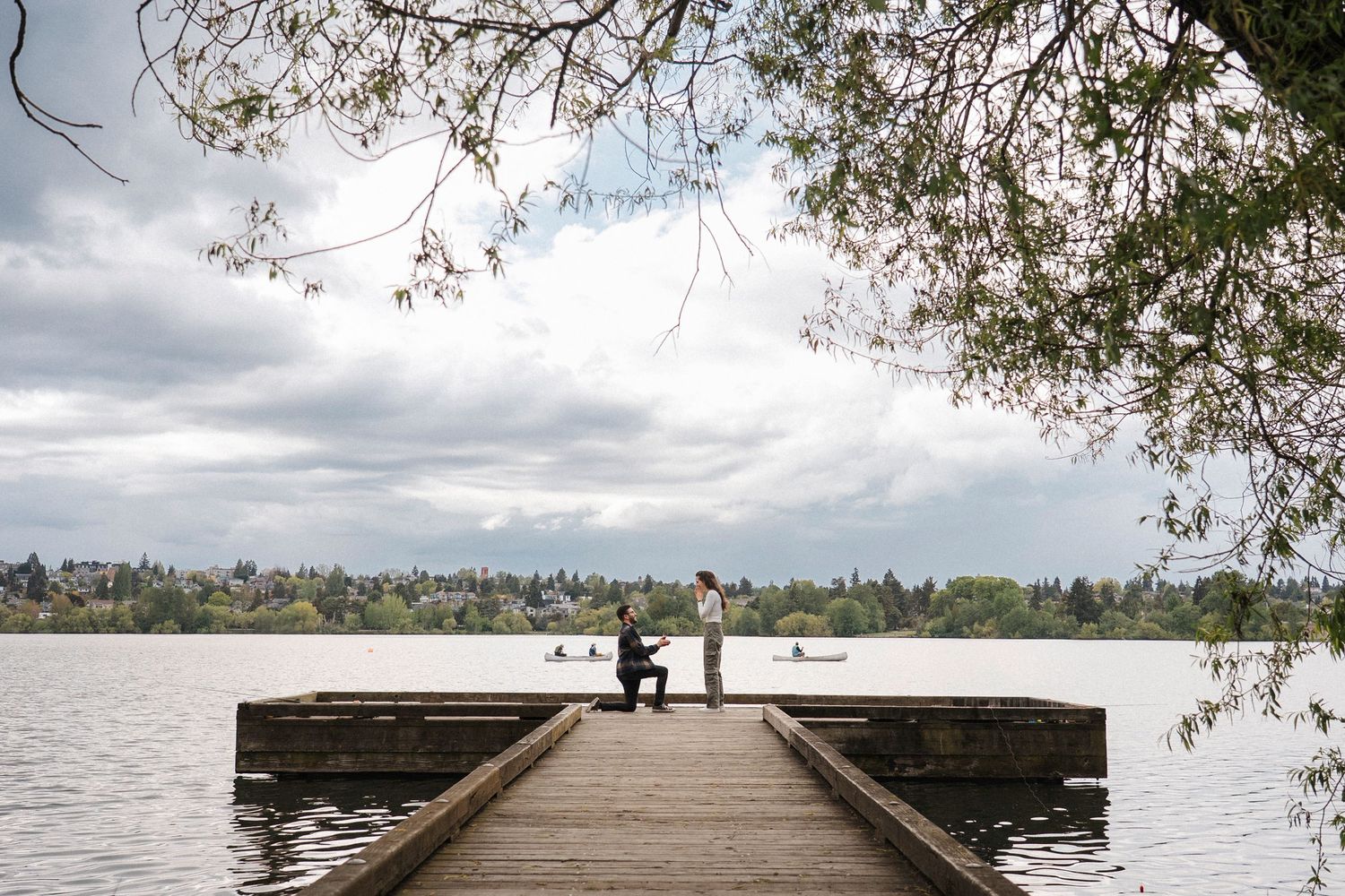 A solitary figure sits on a wooden pier overlooking a calm lake with tree branches framing the cloudy sky.