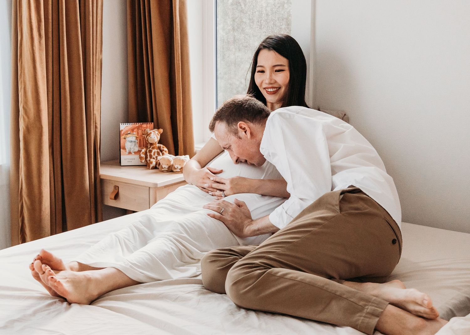 Couple sharing a joyful moment together while relaxing on bed at home with brown curtains in background.
