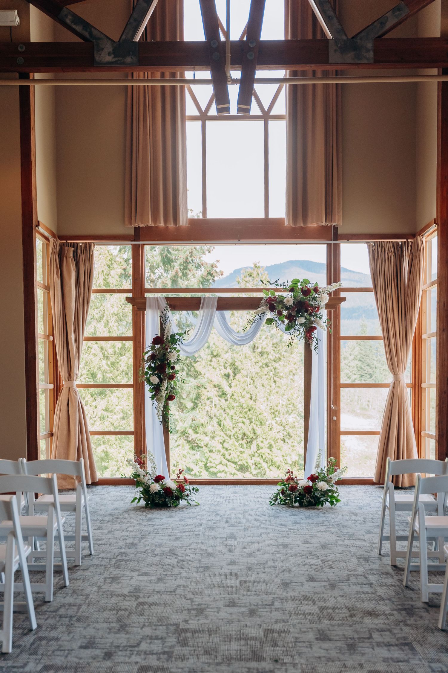 Modern wedding venue interior with large windows, wooden beams, and white chairs set up for ceremony.