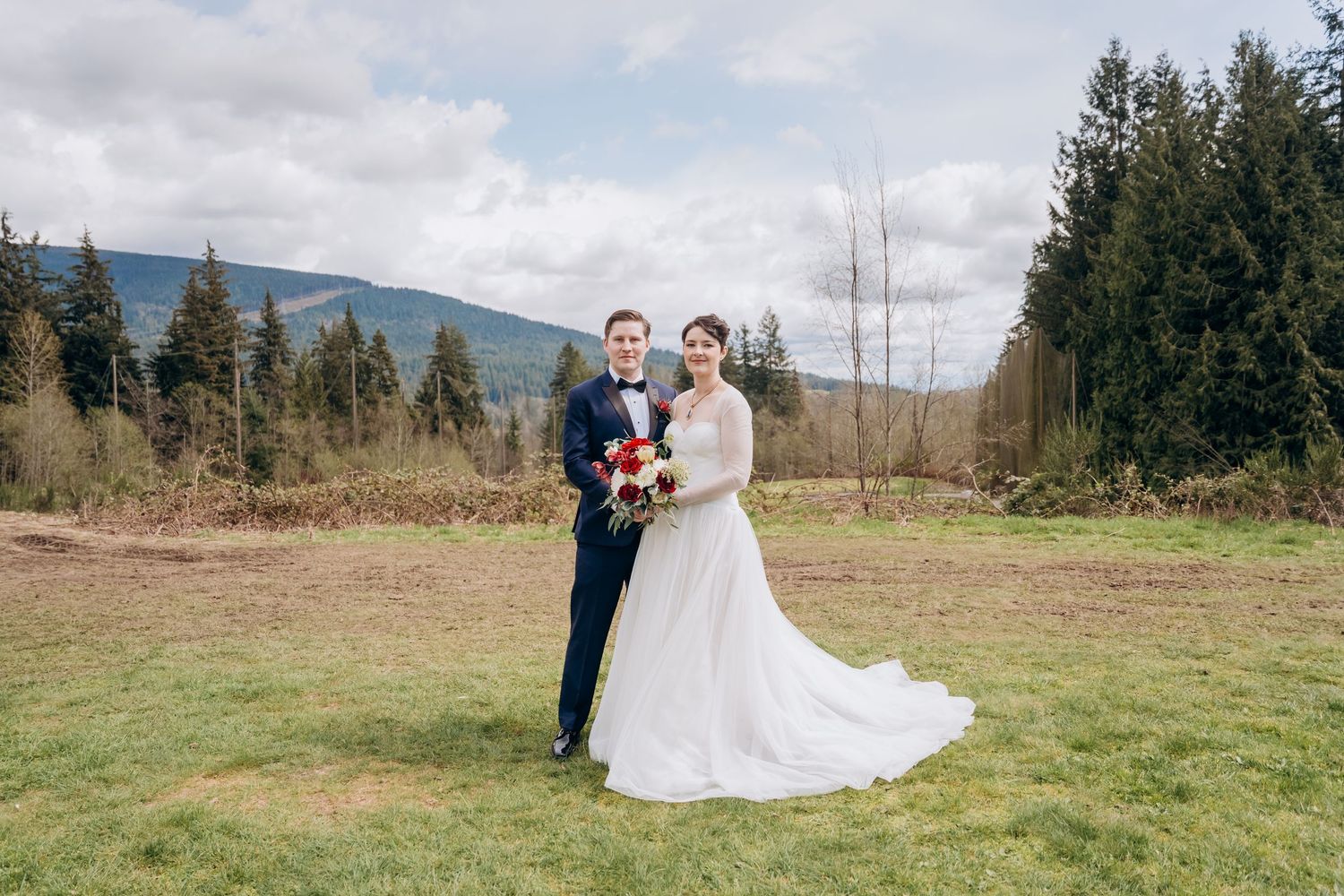 Wedding couple in an open field with mountain landscape and cloudy sky backdrop.