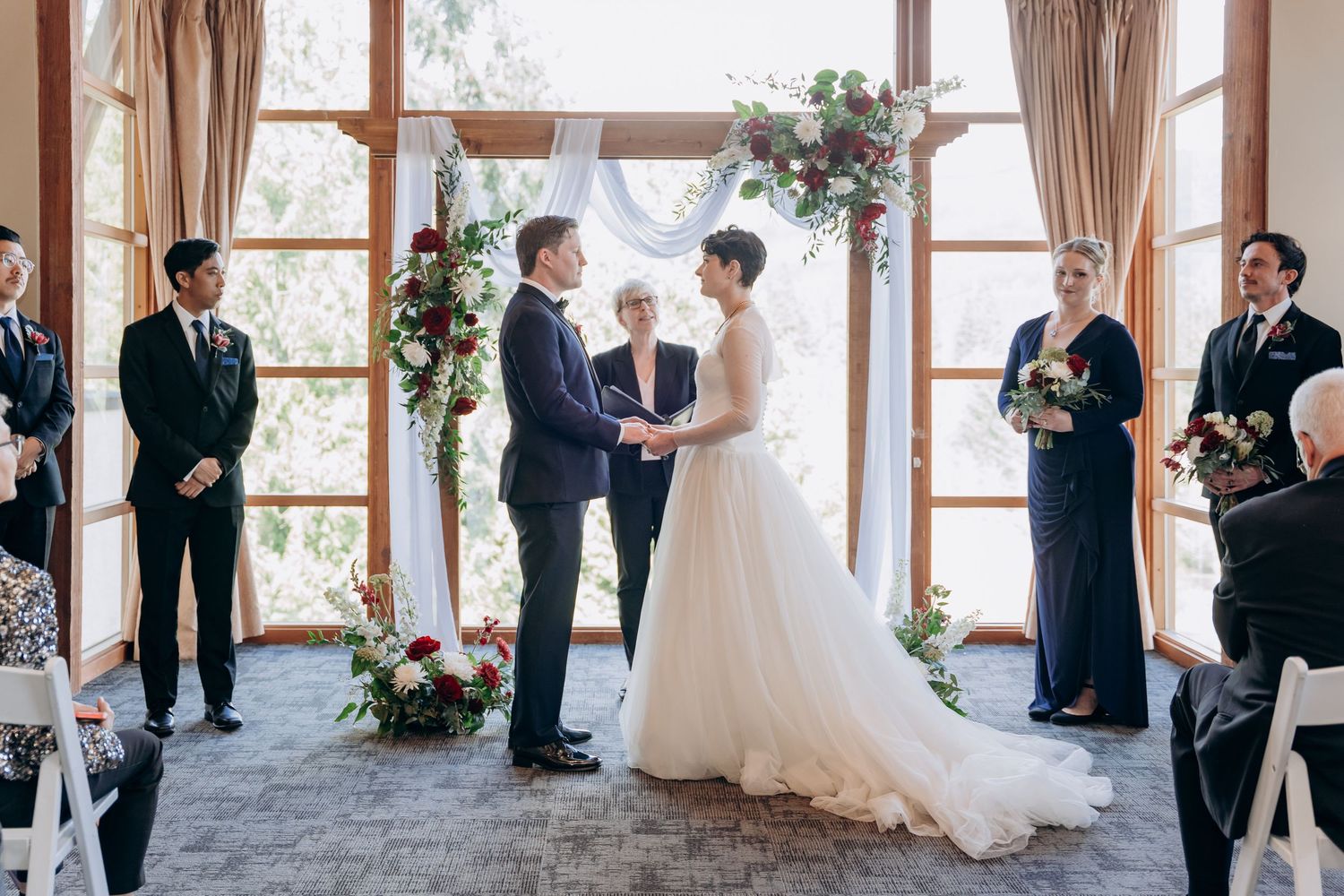 Elegant indoor wedding ceremony with guests gathered in a room featuring large windows and natural light.