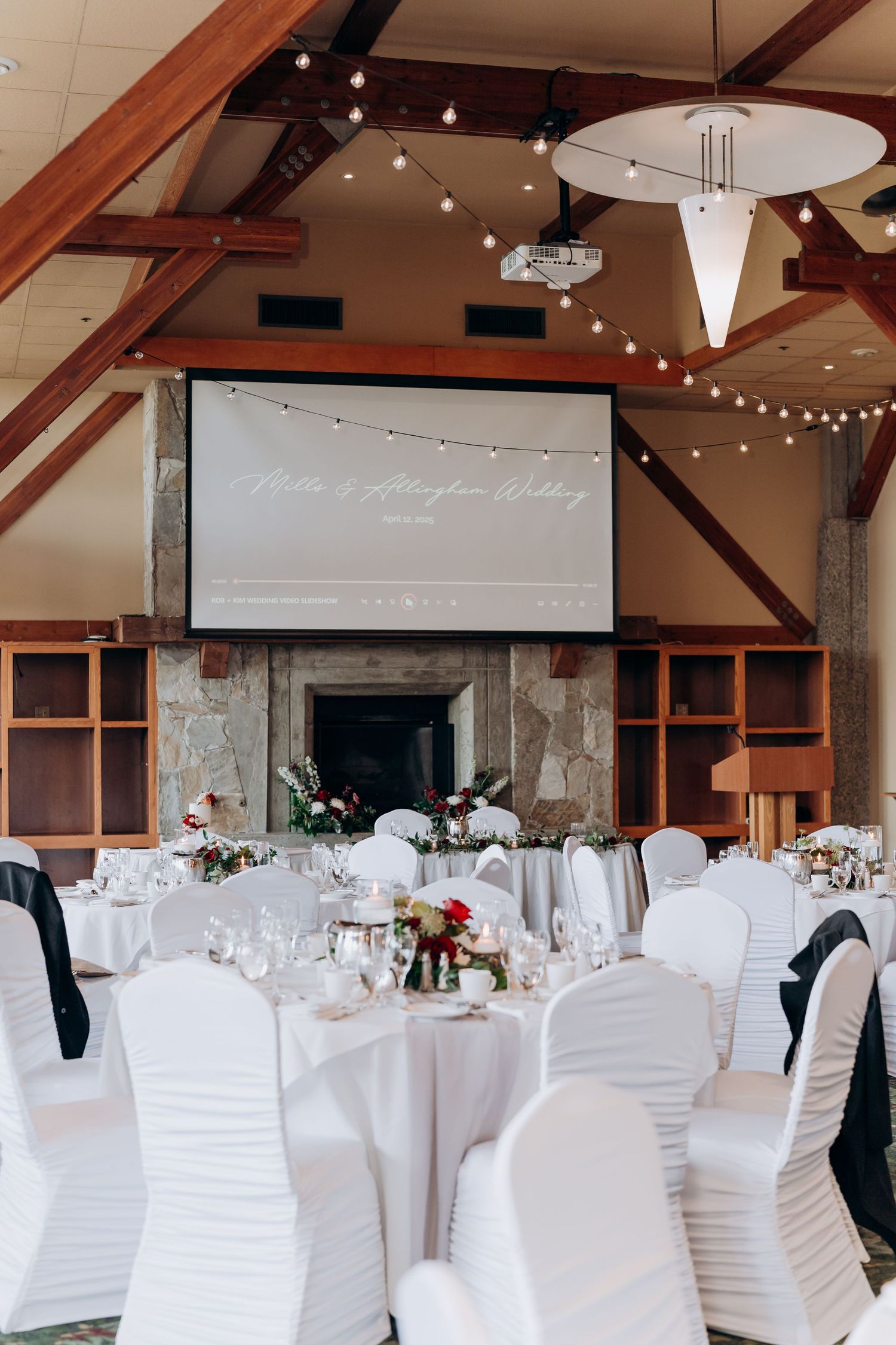 Wedding reception hall decorated with white chair covers and elegant table settings under wooden beams.