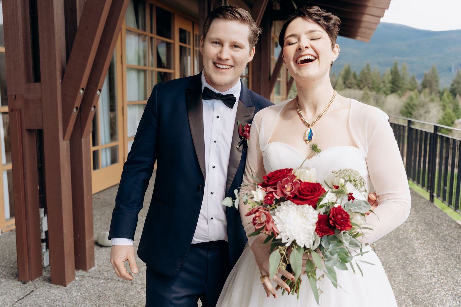 Joyful wedding couple laughing together outdoors with red and white floral bouquet.