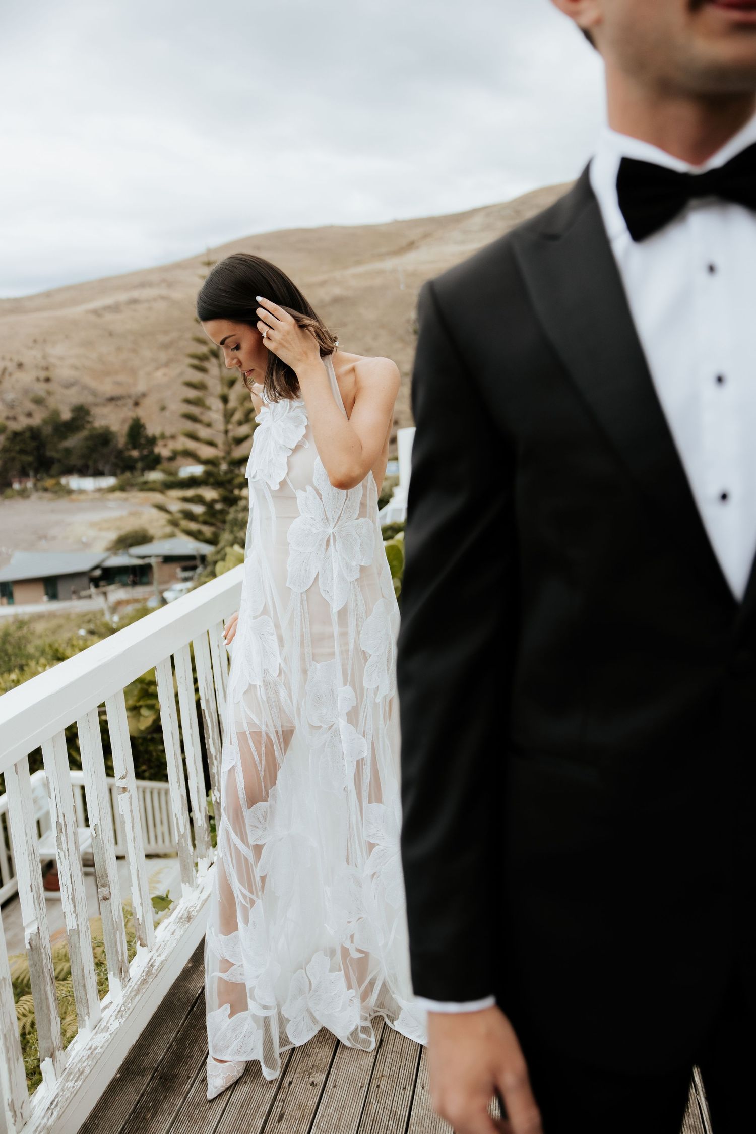 A wedding couple in formal attire standing on a deck with mountain views in the background.