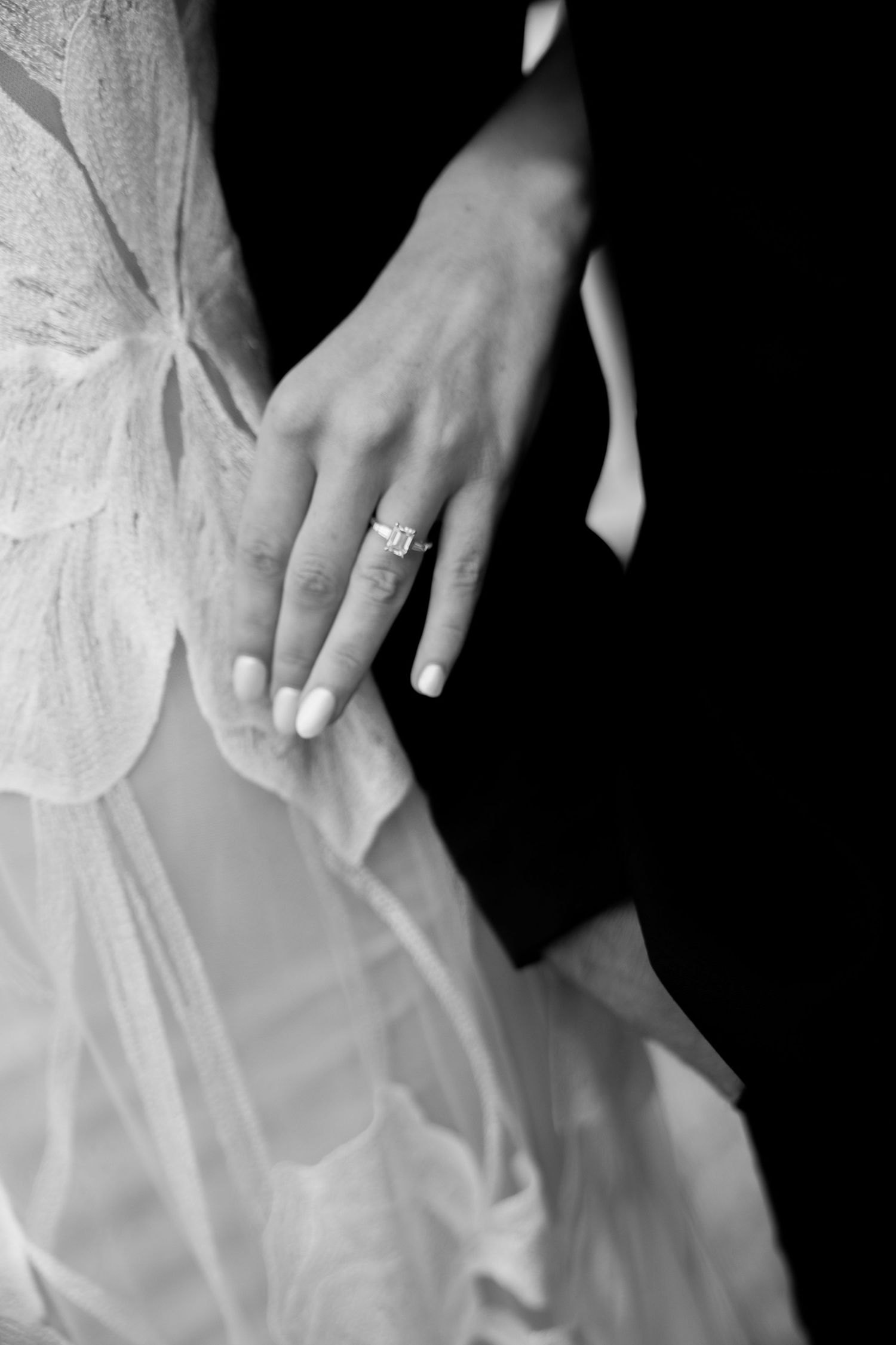Close-up black and white photo of engagement ring on bride's hand against white dress.