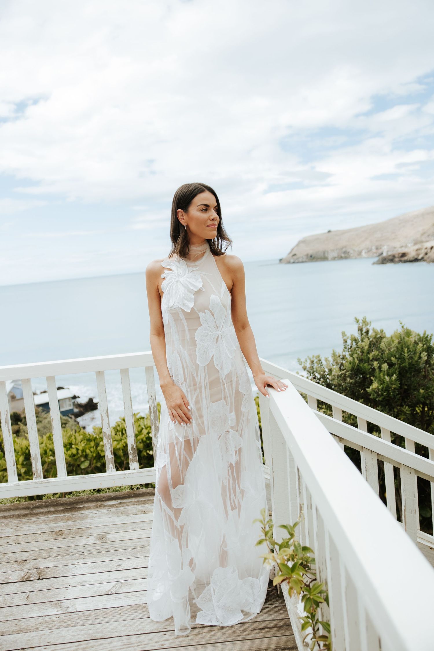 Bride in flowing white dress poses on wooden deck with ocean view.