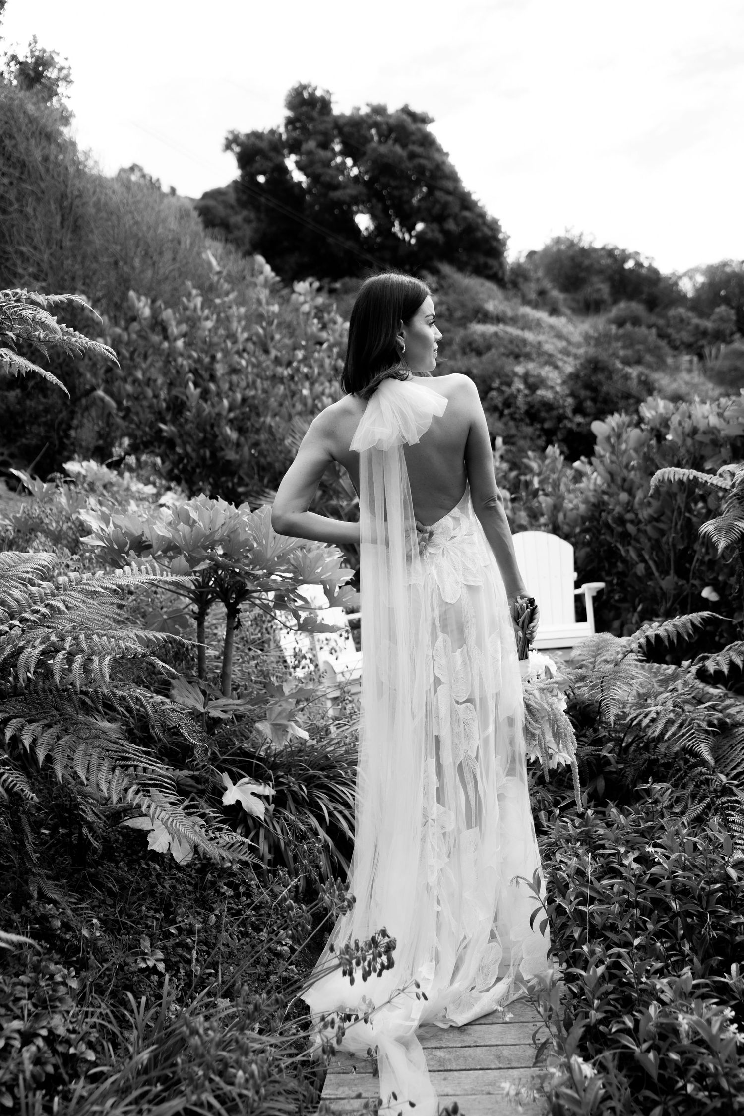 Black and white portrait of bride in flowing dress standing among garden ferns.