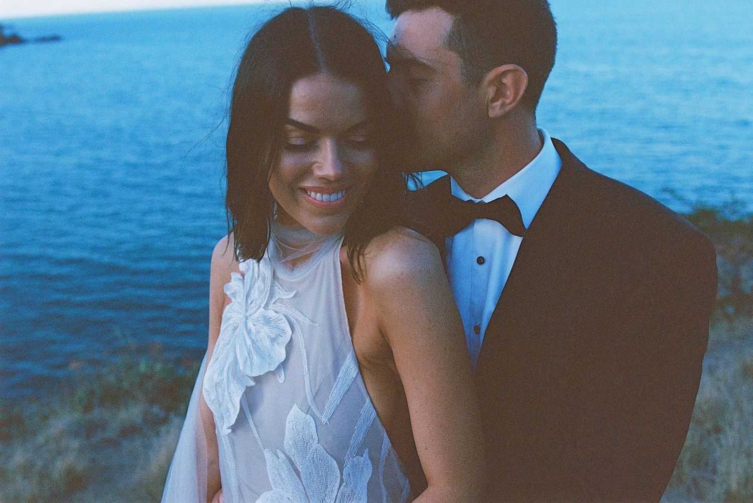 Couple in formal wedding attire embracing by blue ocean on sunny day.